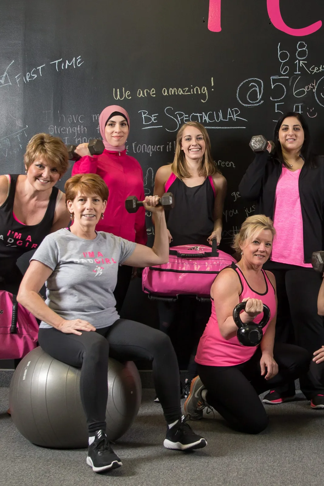 A diverse group of Bad Girl Fitness members, one wearing a pink hijab, holding dumbbells, kettlebells and a pink sandbag and smiling in front of the studio chalkboard wall reading I CAN and Be Spectacular